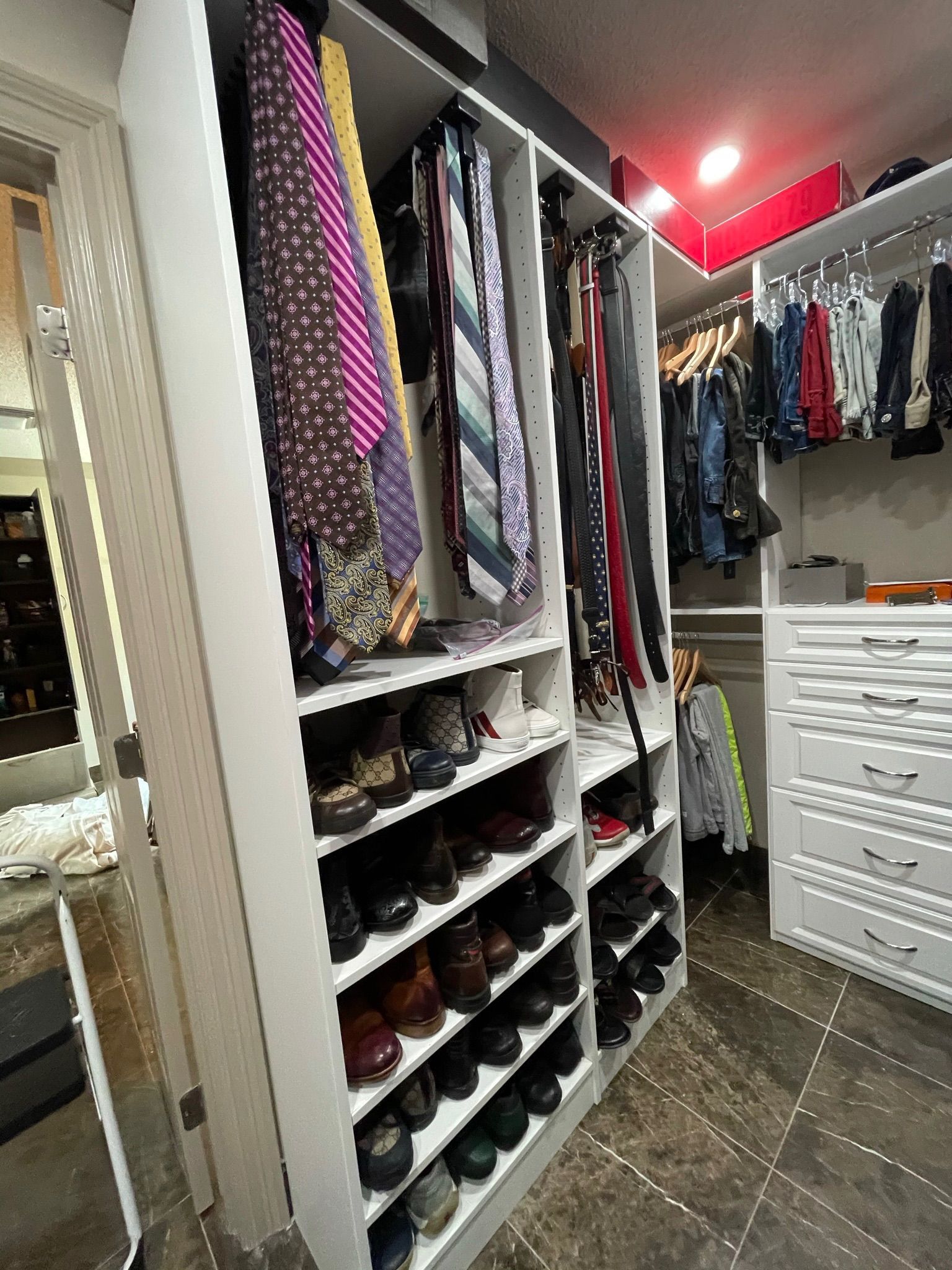 Walk-in closet with shelves of shoes, ties, and hanging clothes; white cabinetry on stone floor.
