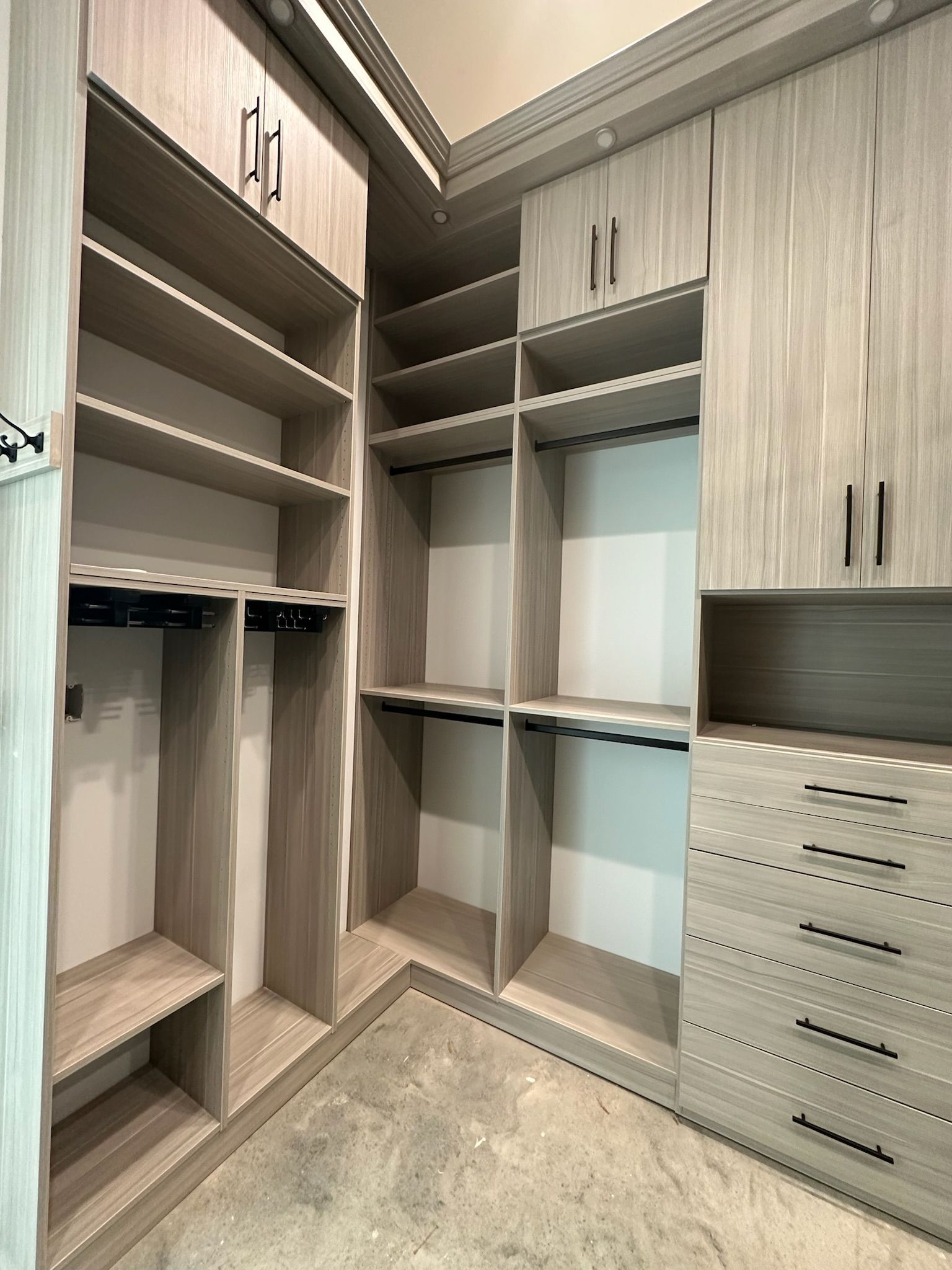 Walk-in closet with light gray wood cabinets, shelves, drawers, and hanging rods. The floor is concrete.