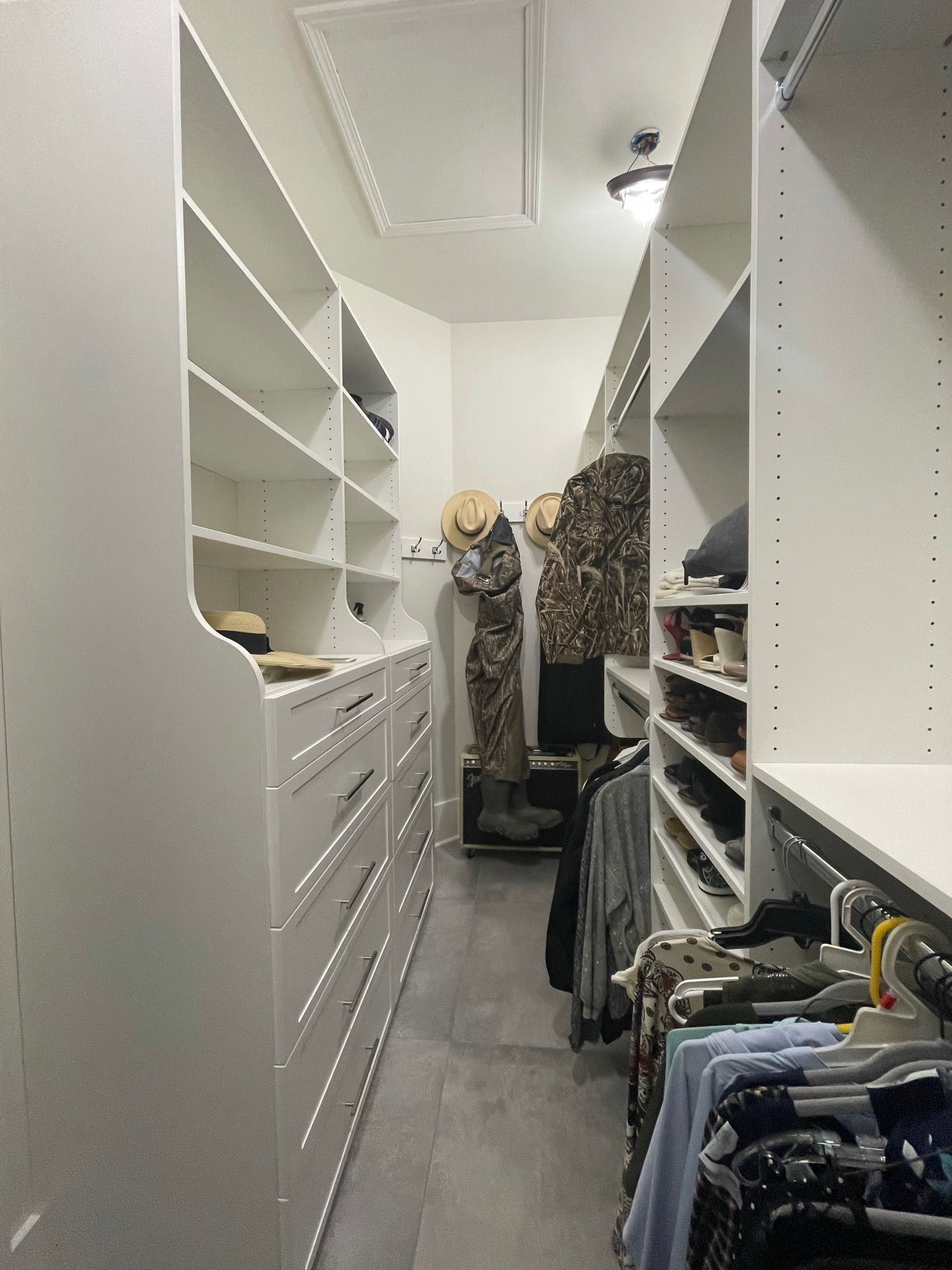 White walk-in closet with shelves, drawers, and hanging clothes. Gray flooring and a light fixture are visible.