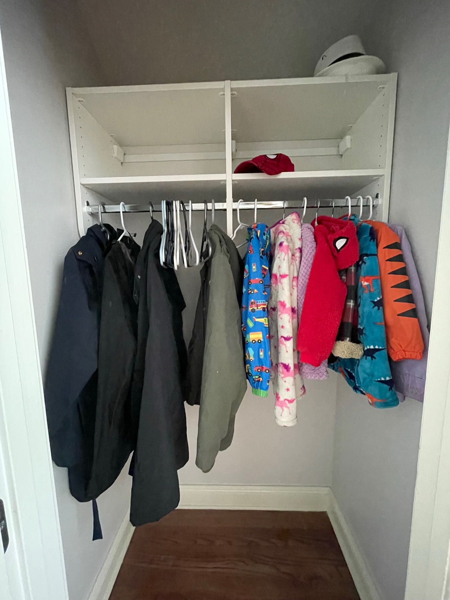 A small closet with coats and clothes hanging from a rod, shelves above. A hat sits on the top shelf.