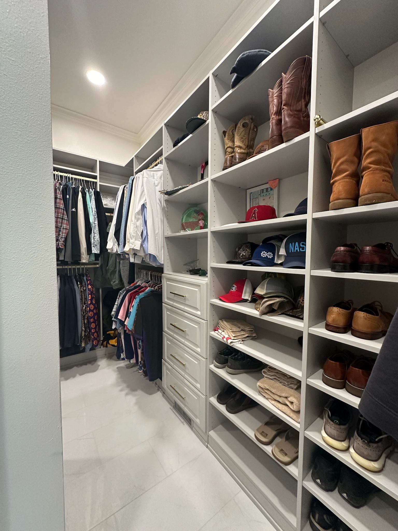 Walk-in closet with white shelves and drawers, filled with shoes, boots, clothes, and hats.