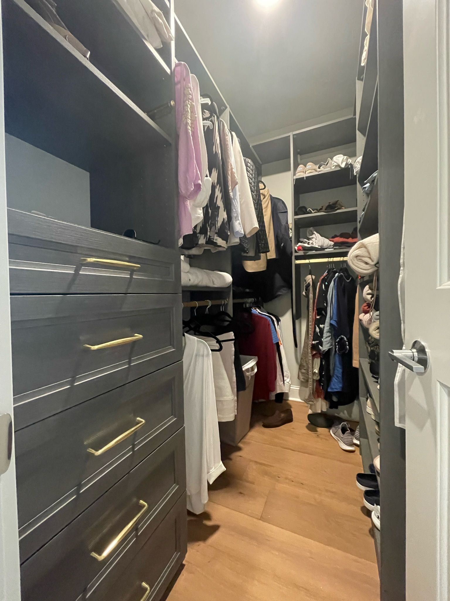 Walk-in closet with dark gray built-ins, clothes hanging on racks, and herringbone wood floor.