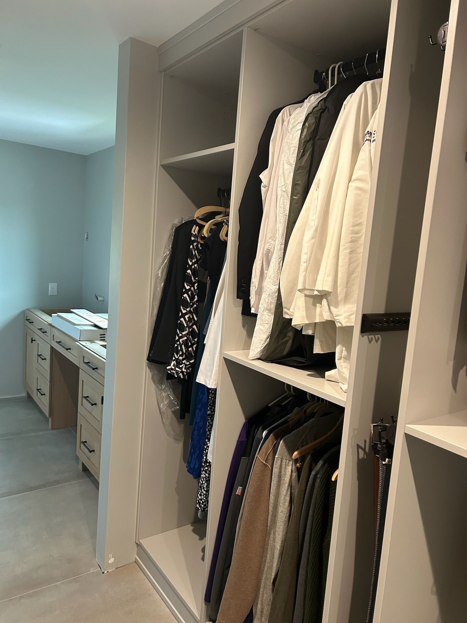 A walk-in closet with hanging clothes in shades of black, white, and gray, with a desk visible in the background.