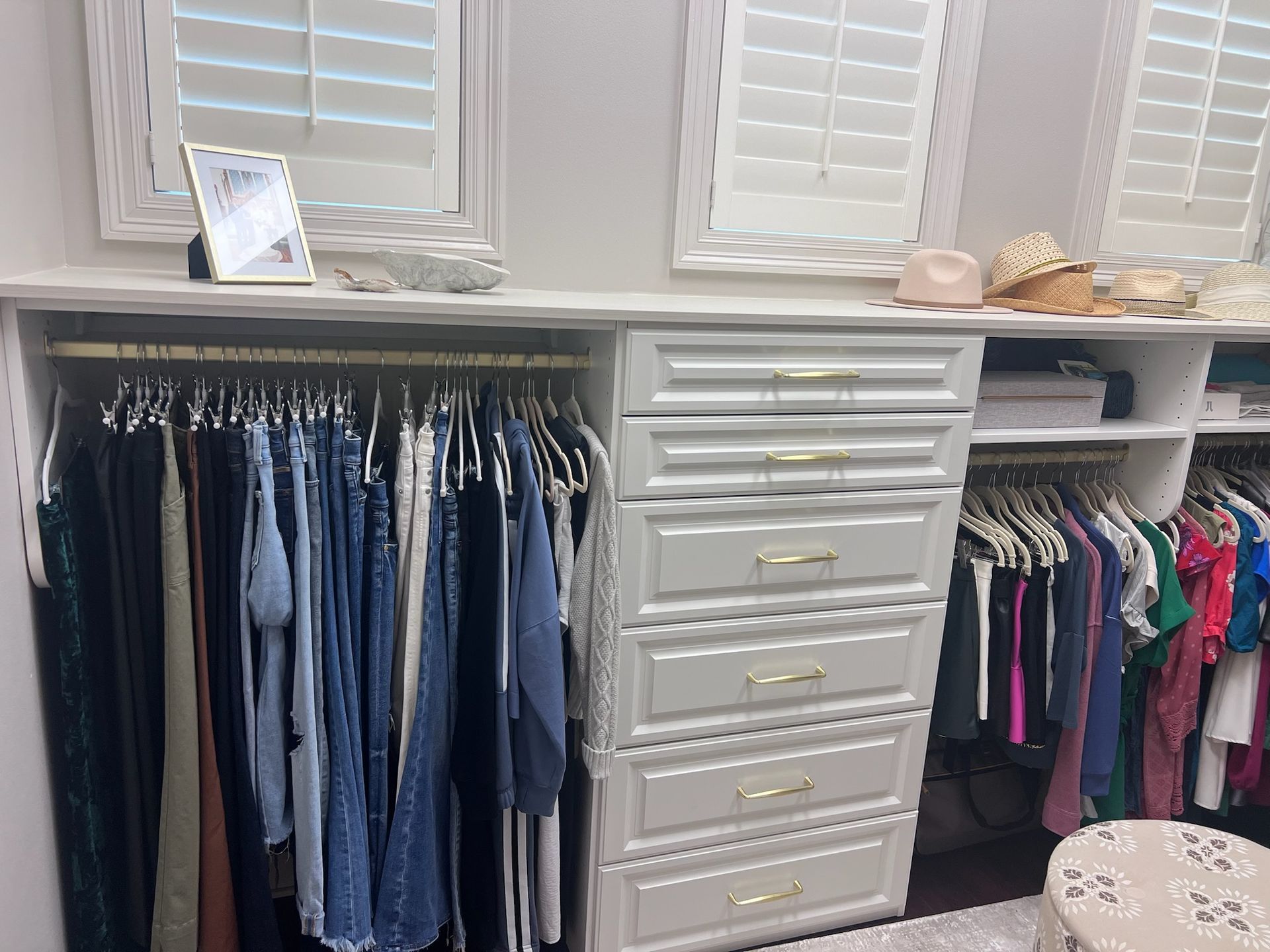 Closet with clothes on hangers and a chest of drawers. Windows and shelves above.