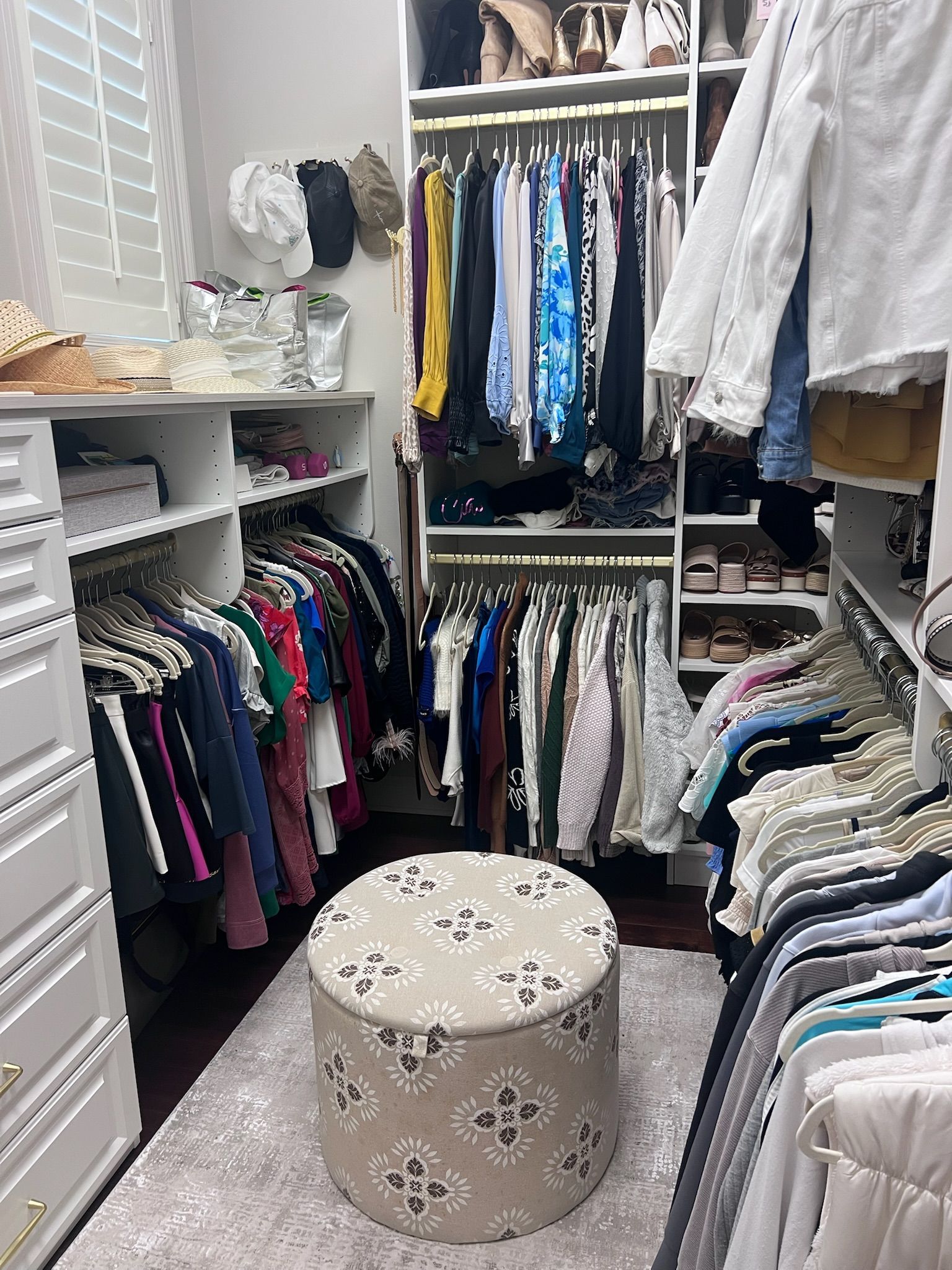 Walk-in closet with clothes on racks, shelves with shoes, a pouf in the center.