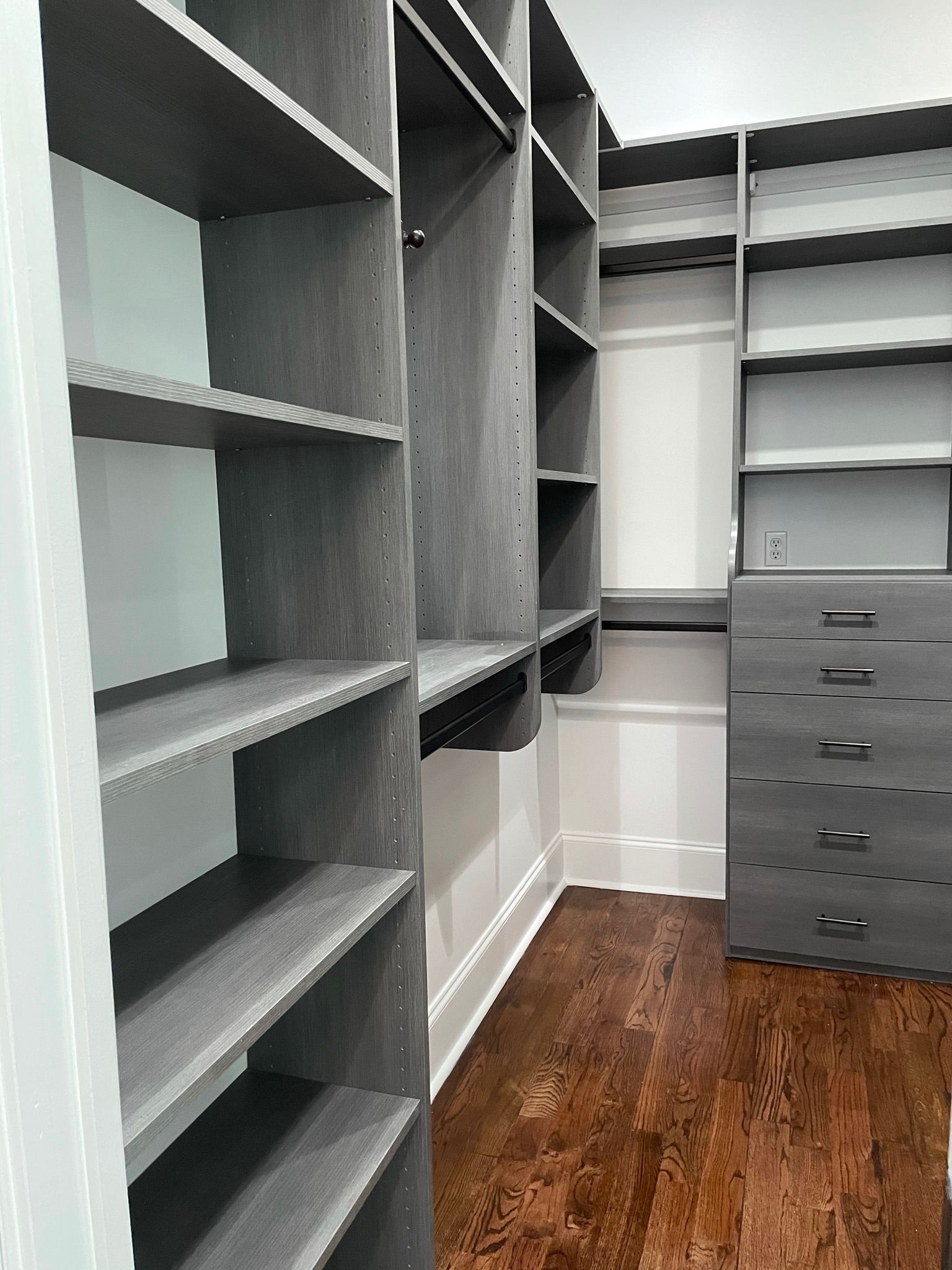 Walk-in closet with dark gray shelving and drawers, brown wood floor, and white walls.