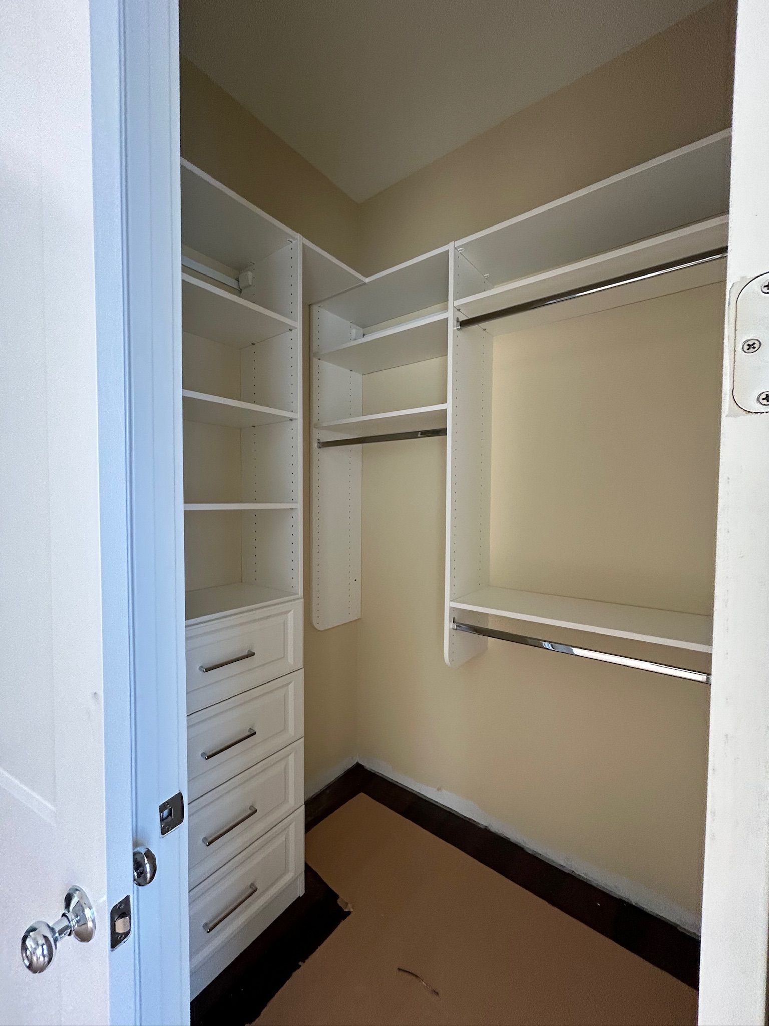 Empty walk-in closet with white shelving and drawers. Dark floor trim and tan walls. Blue door frame.