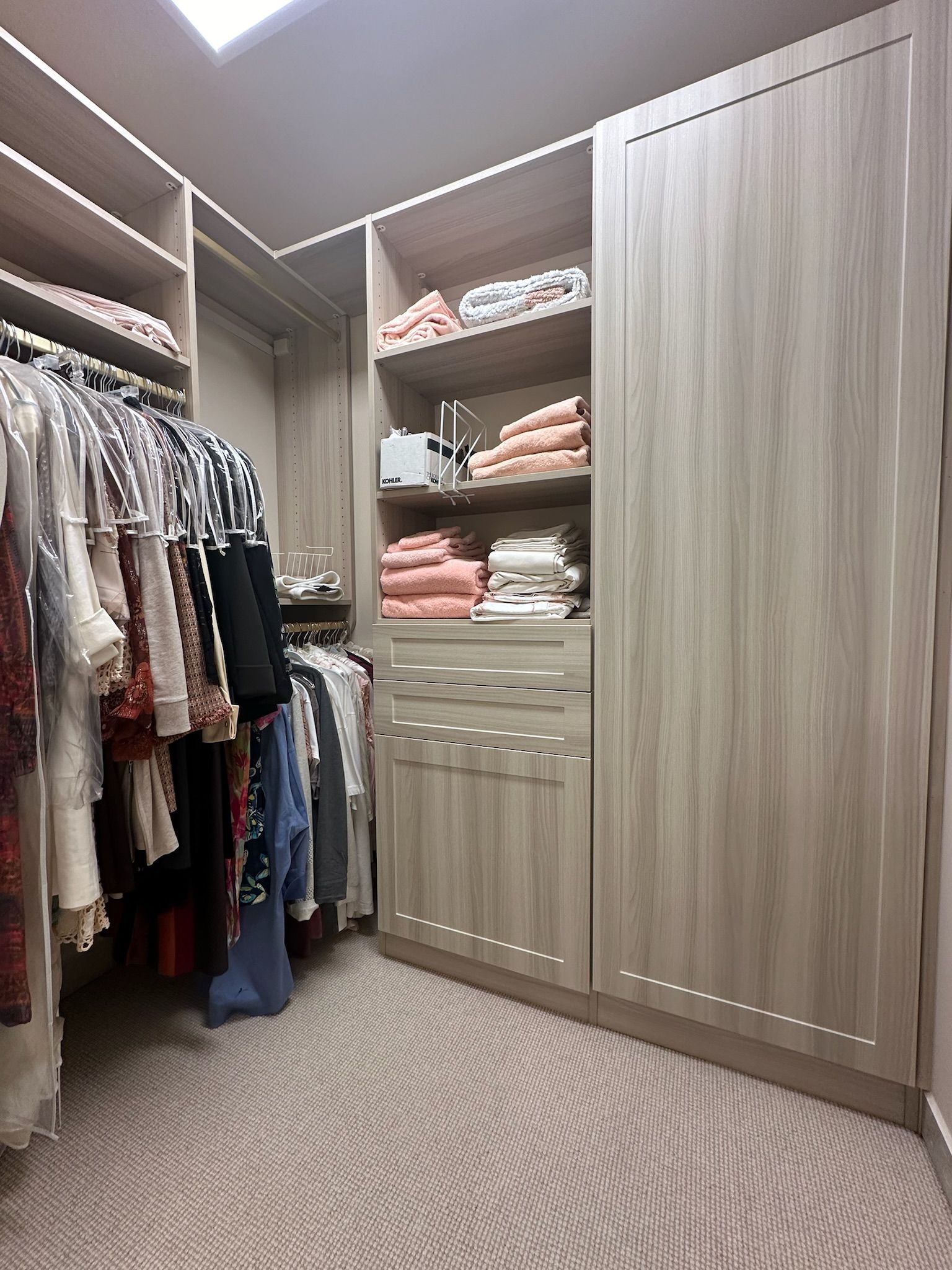 Walk-in closet with light wood shelving and hanging clothes, plus stacks of folded towels and a patterned carpet.