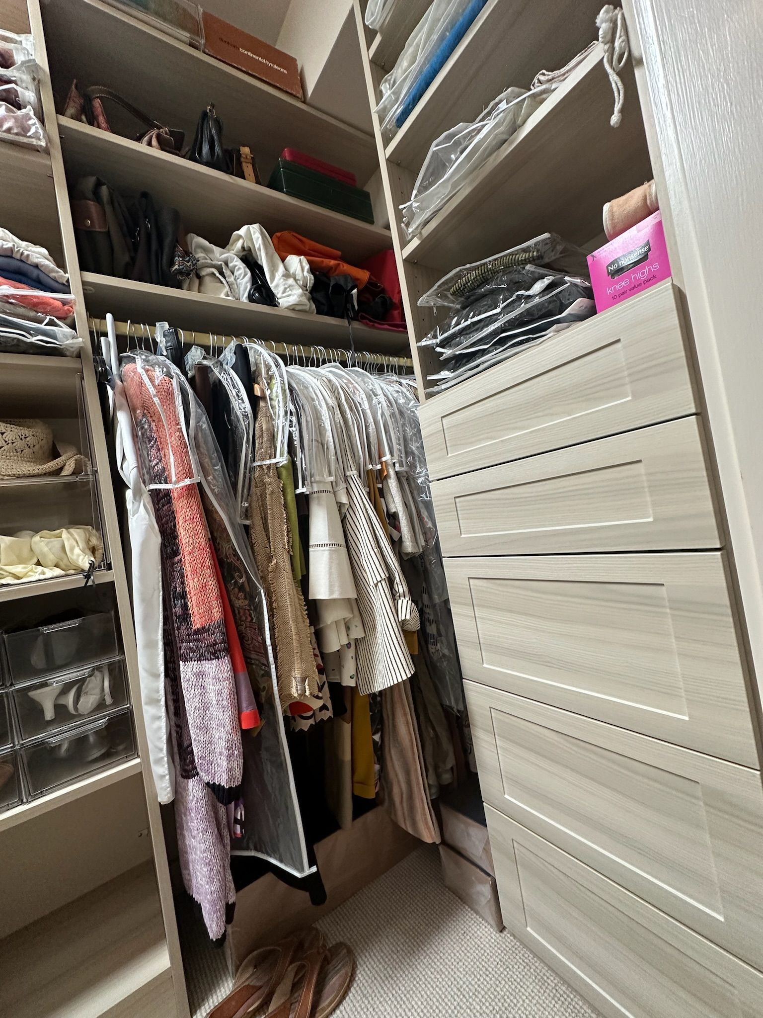 A walk-in closet with clothes hanging on a rod, shelves with bags, and a set of drawers.