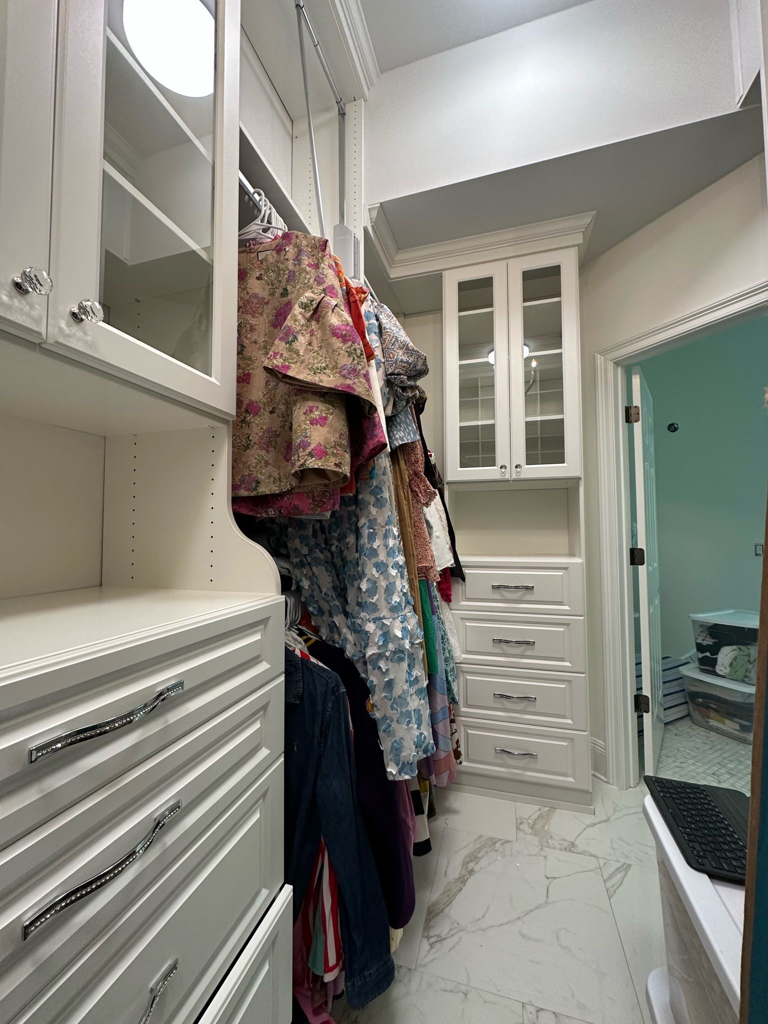 Walk-in closet with white cabinets, drawers, and hanging clothes. Marble-like flooring, a glass-paned cabinet, and doorway.
