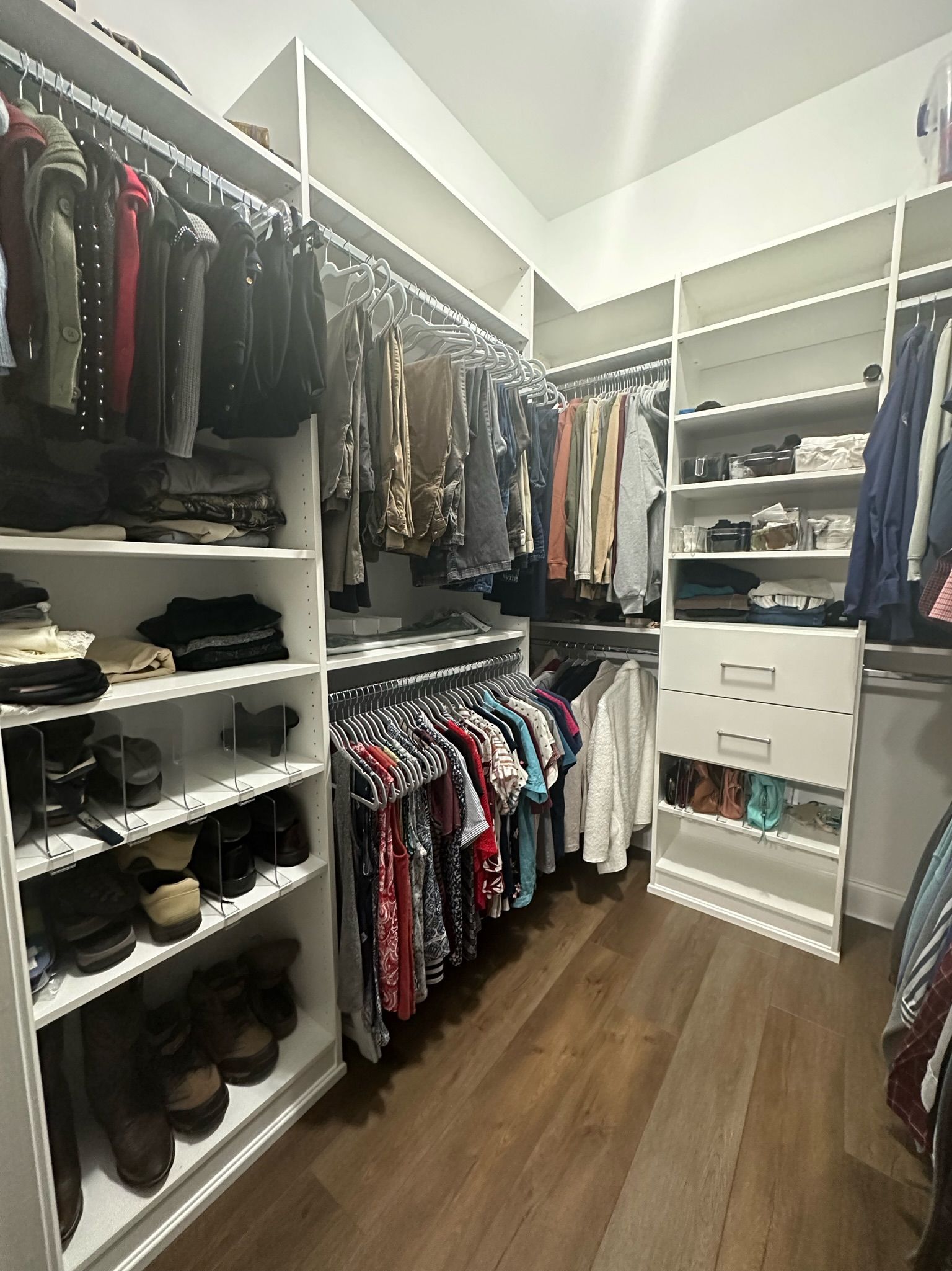 Walk-in closet with white shelves and hanging rods, brown wood floor, and various clothes and shoes.