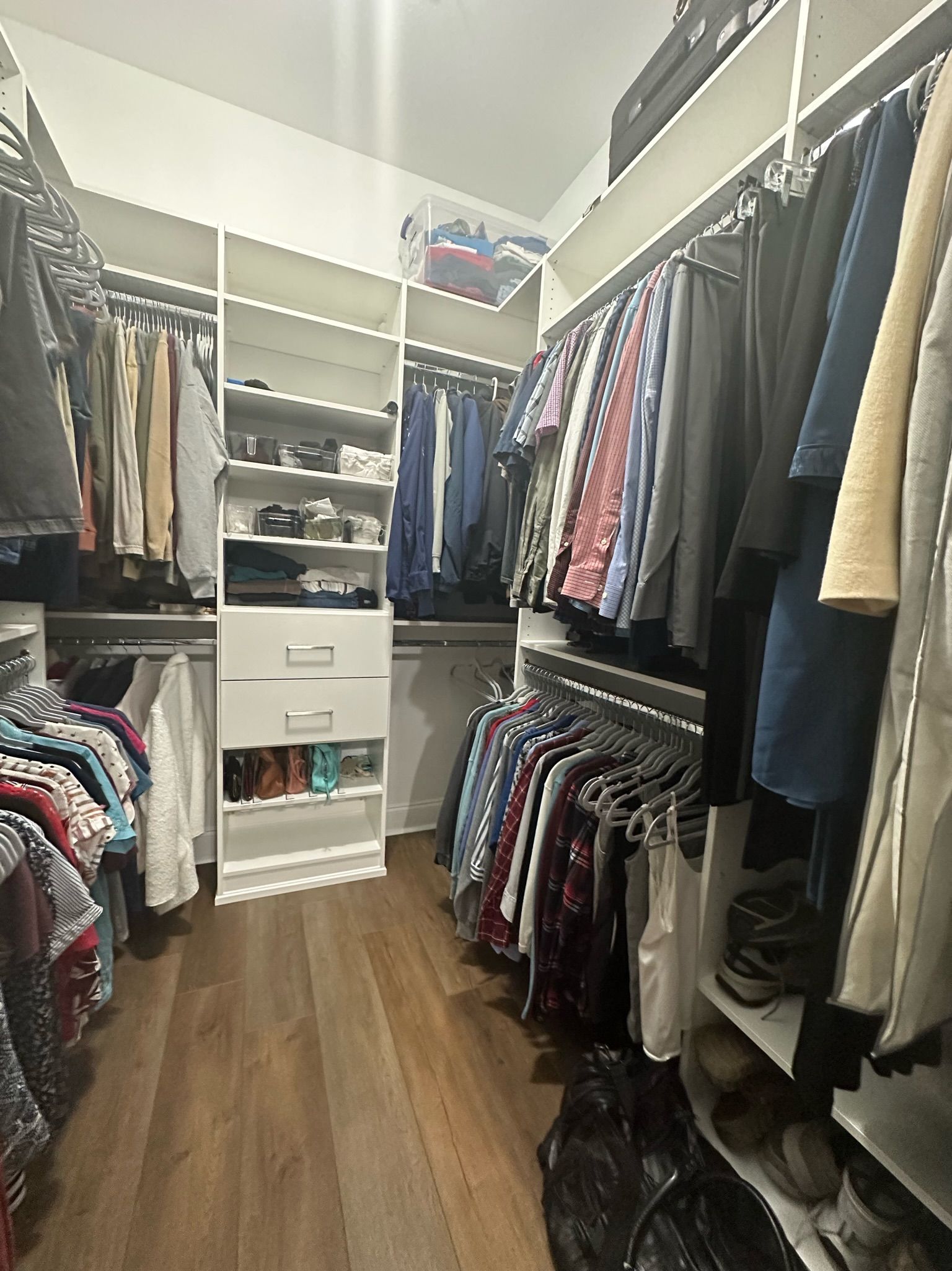 Walk-in closet with white shelves and rods filled with clothes. Wooden floor, overhead light.