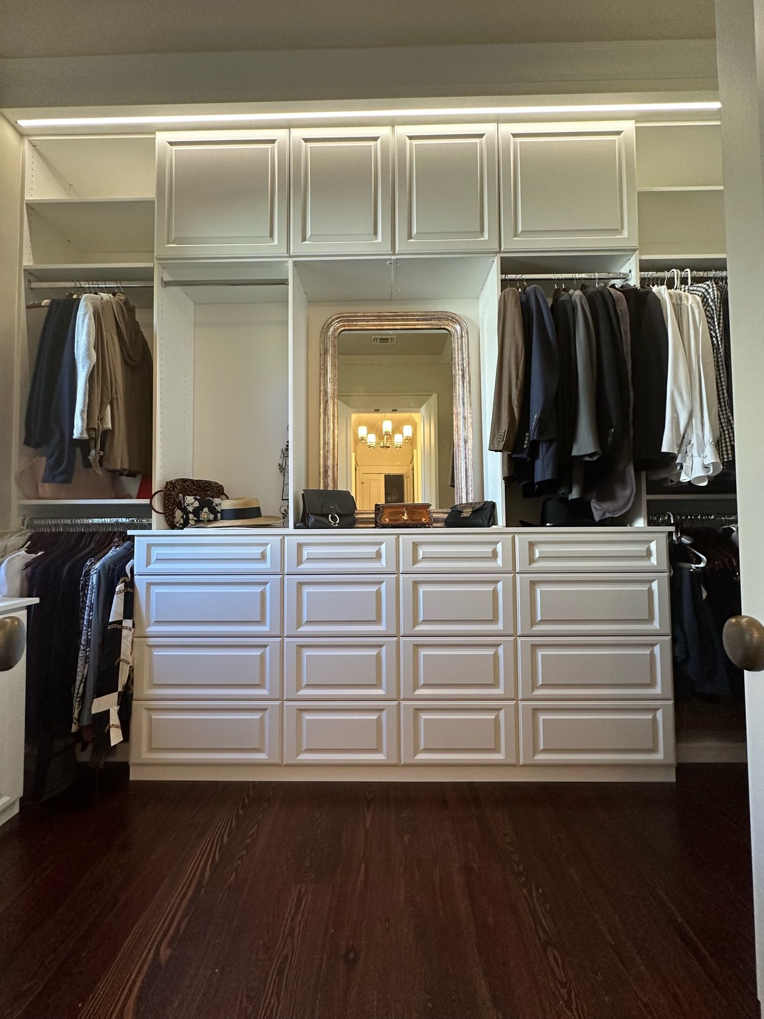 Walk-in closet with white cabinets, drawers, and hanging clothes. Large mirror centered. Dark wood floor.