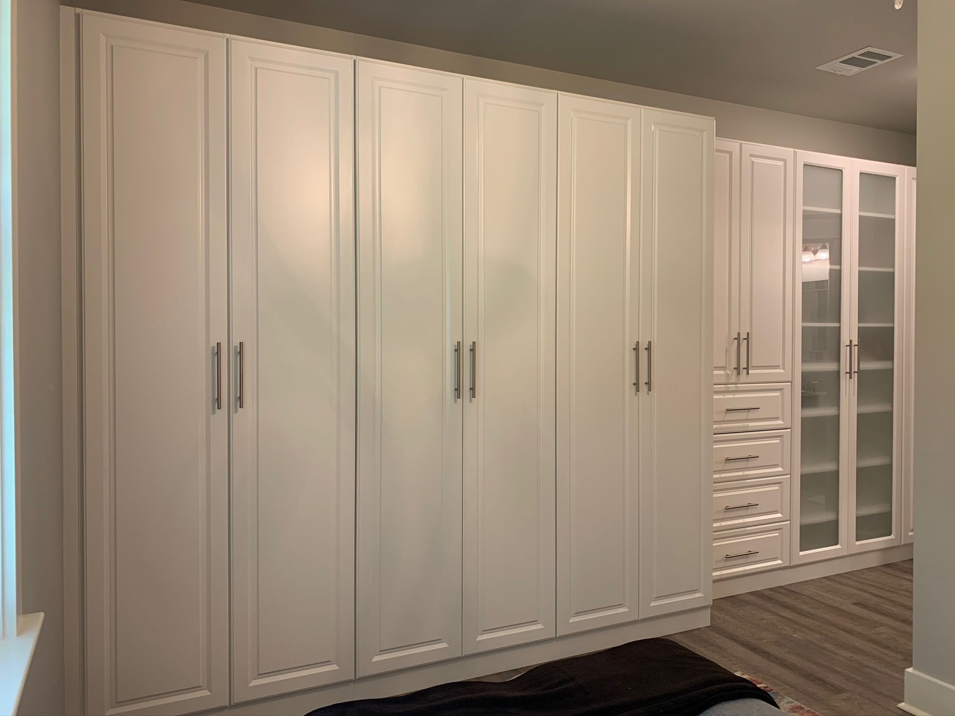 White built-in closet with multiple tall doors and glass-doored sections. Gray floor, neutral walls.