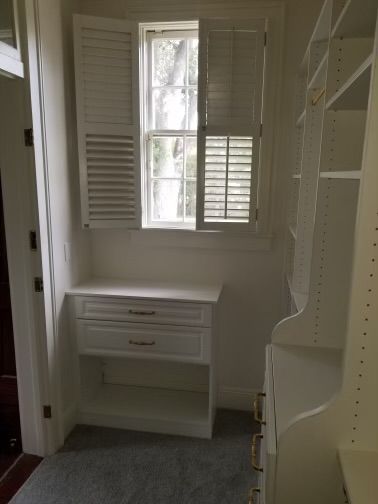 A small white built-in desk with drawers under a window with open shutters in a closet.