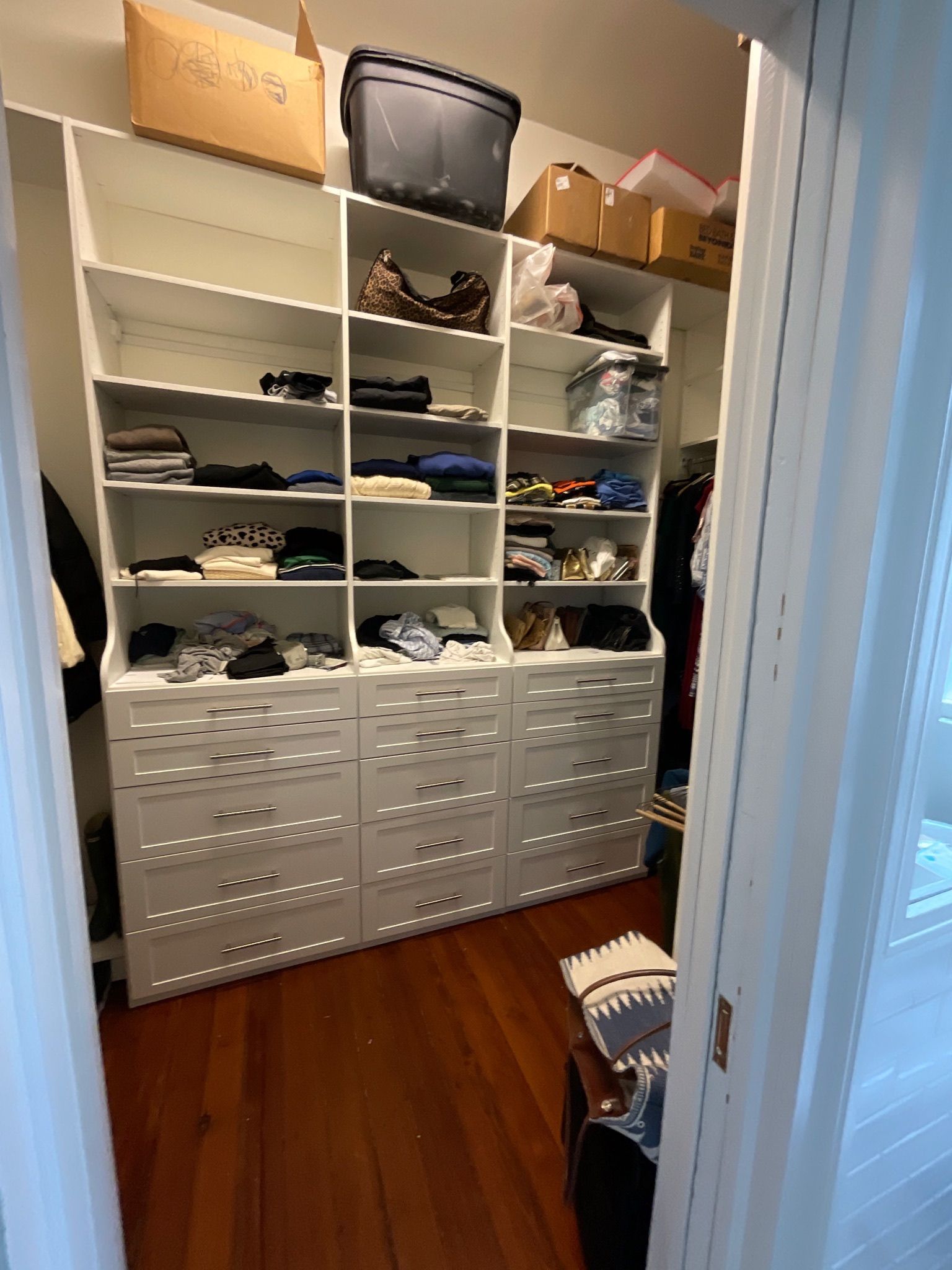 Closet with white shelving and drawers, filled with clothes and storage boxes, hardwood floor.