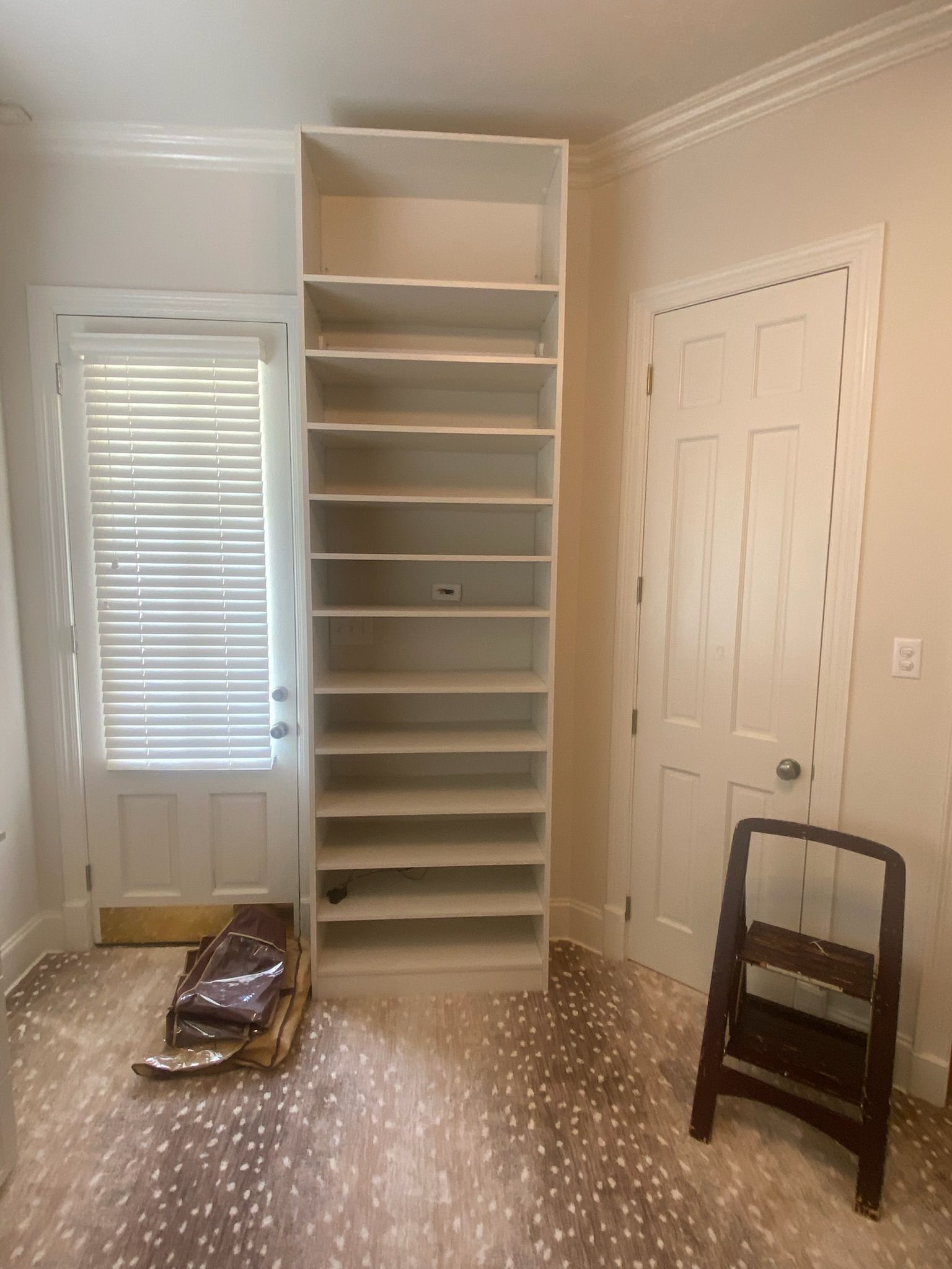 Tall white bookshelf between a door with blinds and a closed door, with a step stool.