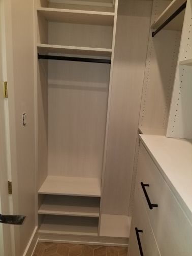 Built-in white closet with shelves, hanging rod, and drawers. Black hardware, tan floor, and light walls.