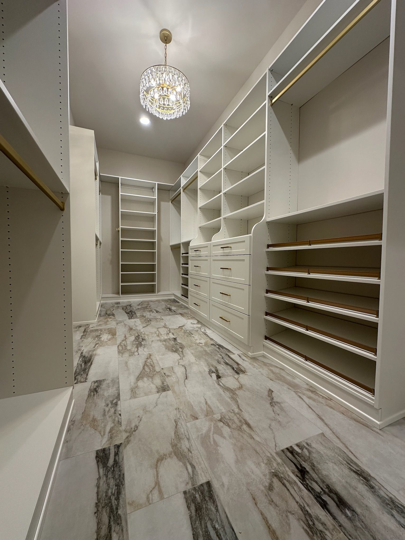 Walk-in closet with white shelves and drawers, marble floor, gold accents, and a crystal chandelier.