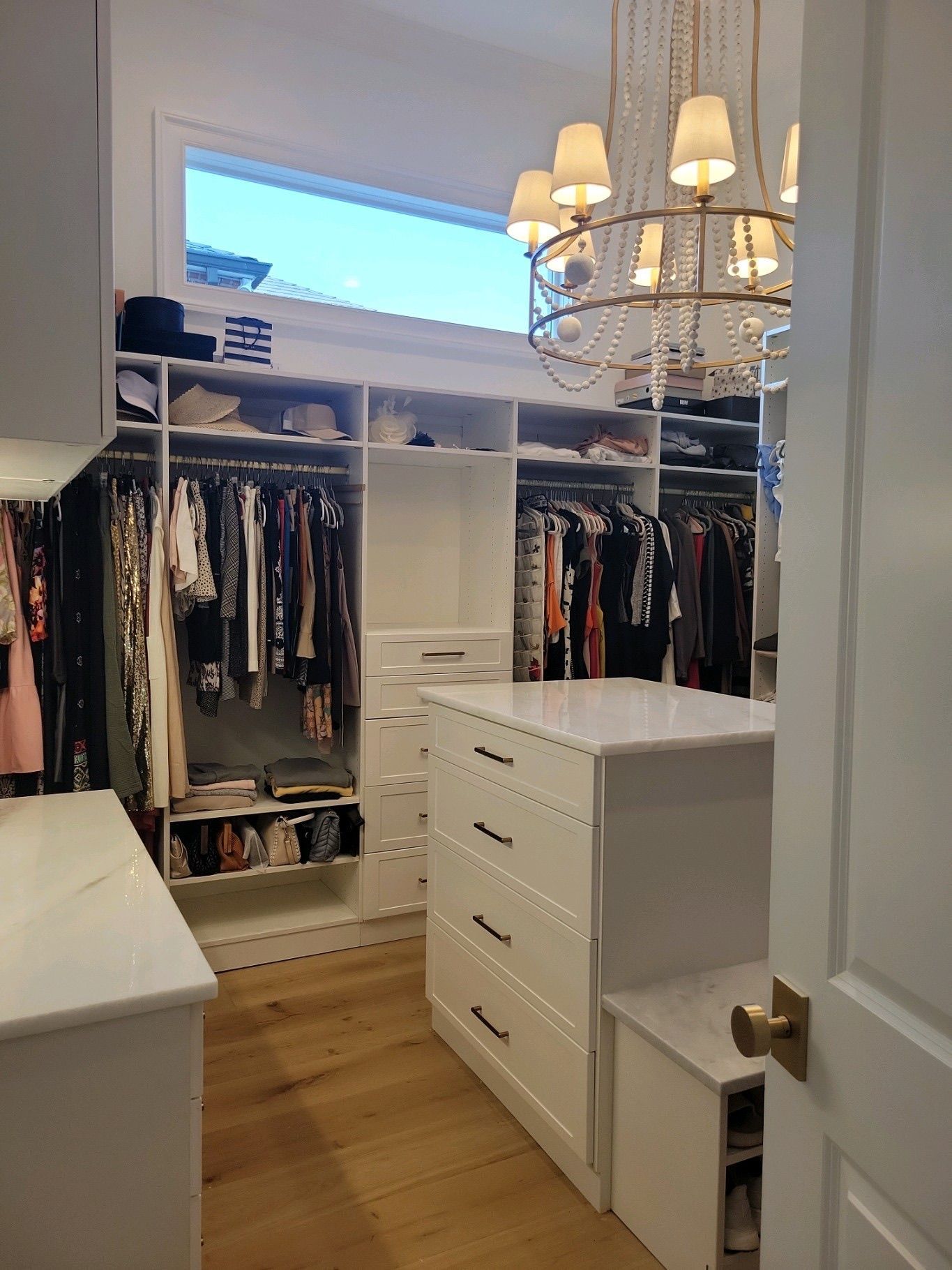 Walk-in closet with white cabinetry, clothing racks, and a central dresser.  A chandelier hangs from the ceiling.