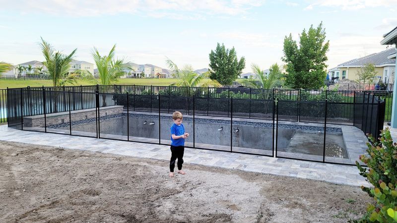 A young boy is standing in front of a fence around a swimming pool.