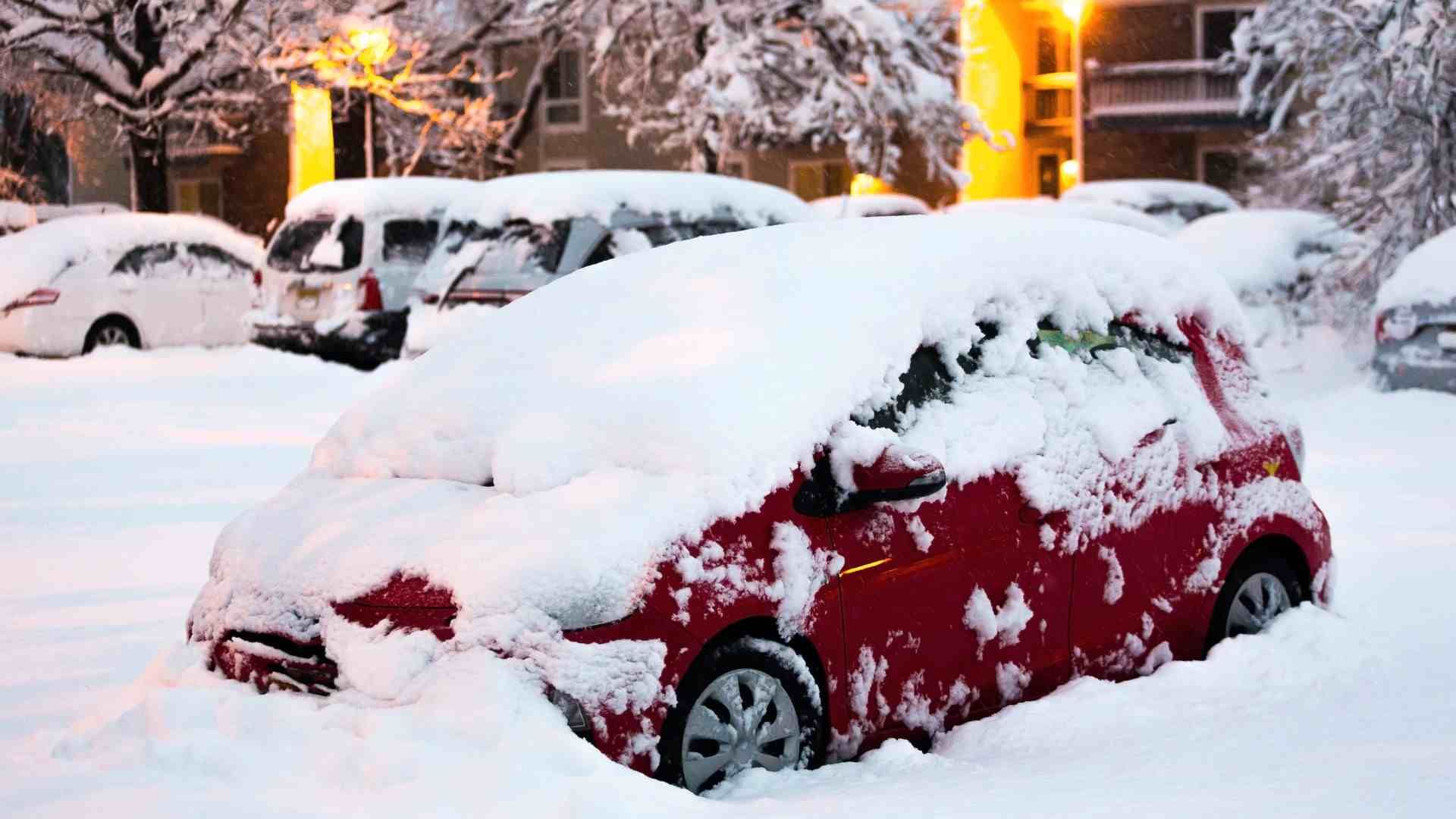 Red car covered in snow, parked in a snow-covered lot. Other cars and buildings are in the background.