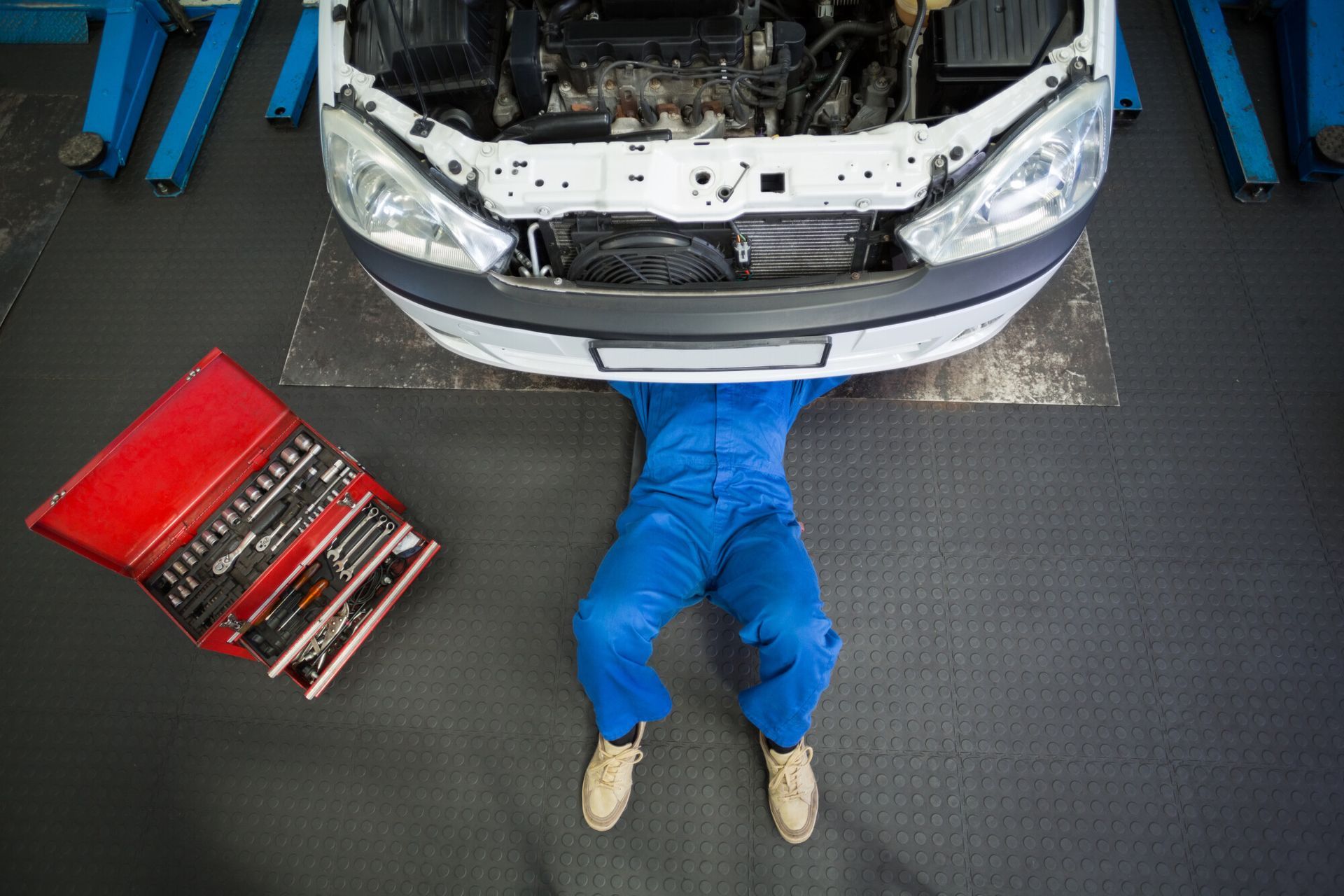 Mechanic in blue jumpsuit working on the underside of a white car in a garage with a toolbox.