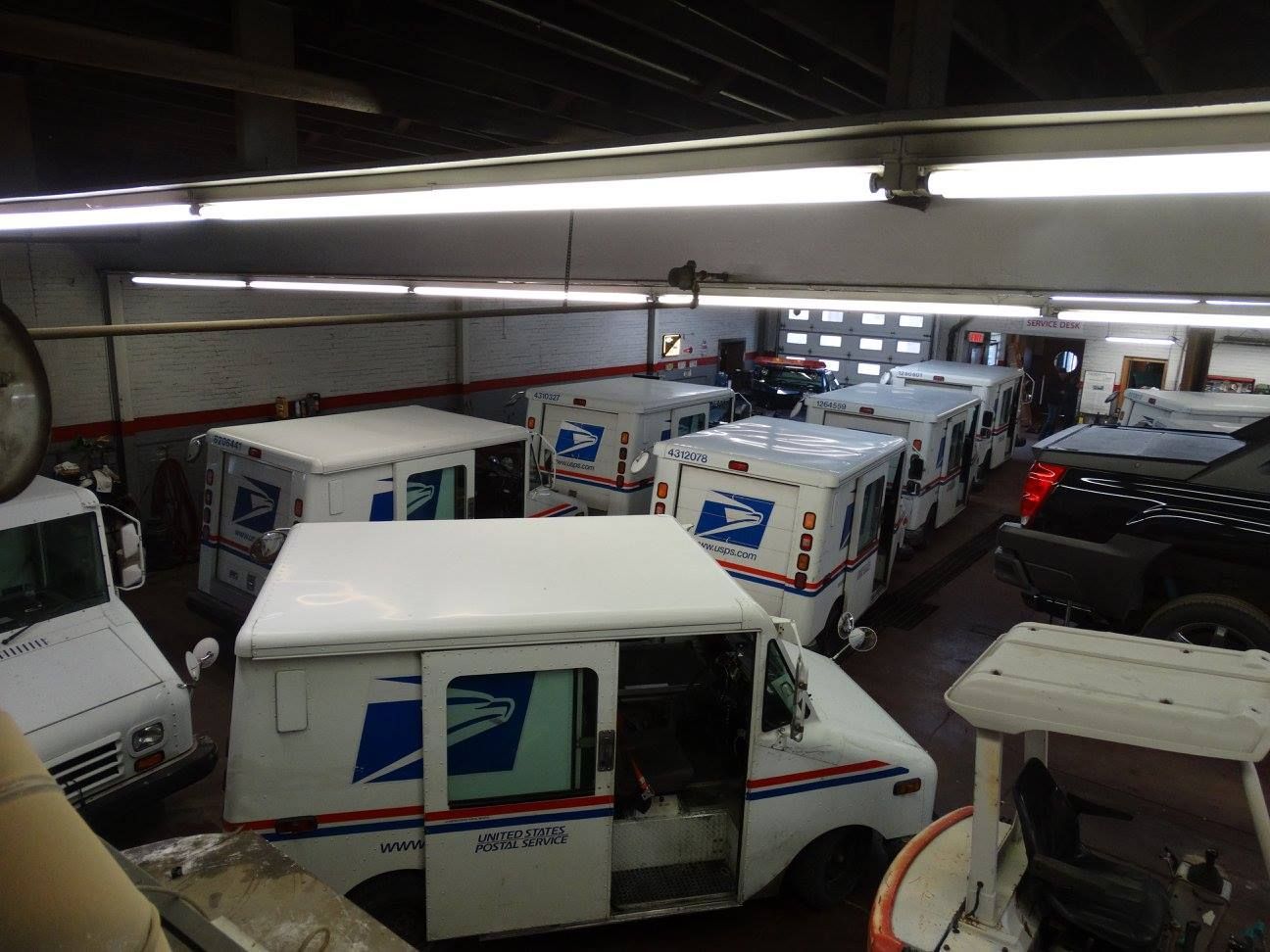Inside a garage, several white USPS mail trucks are parked.