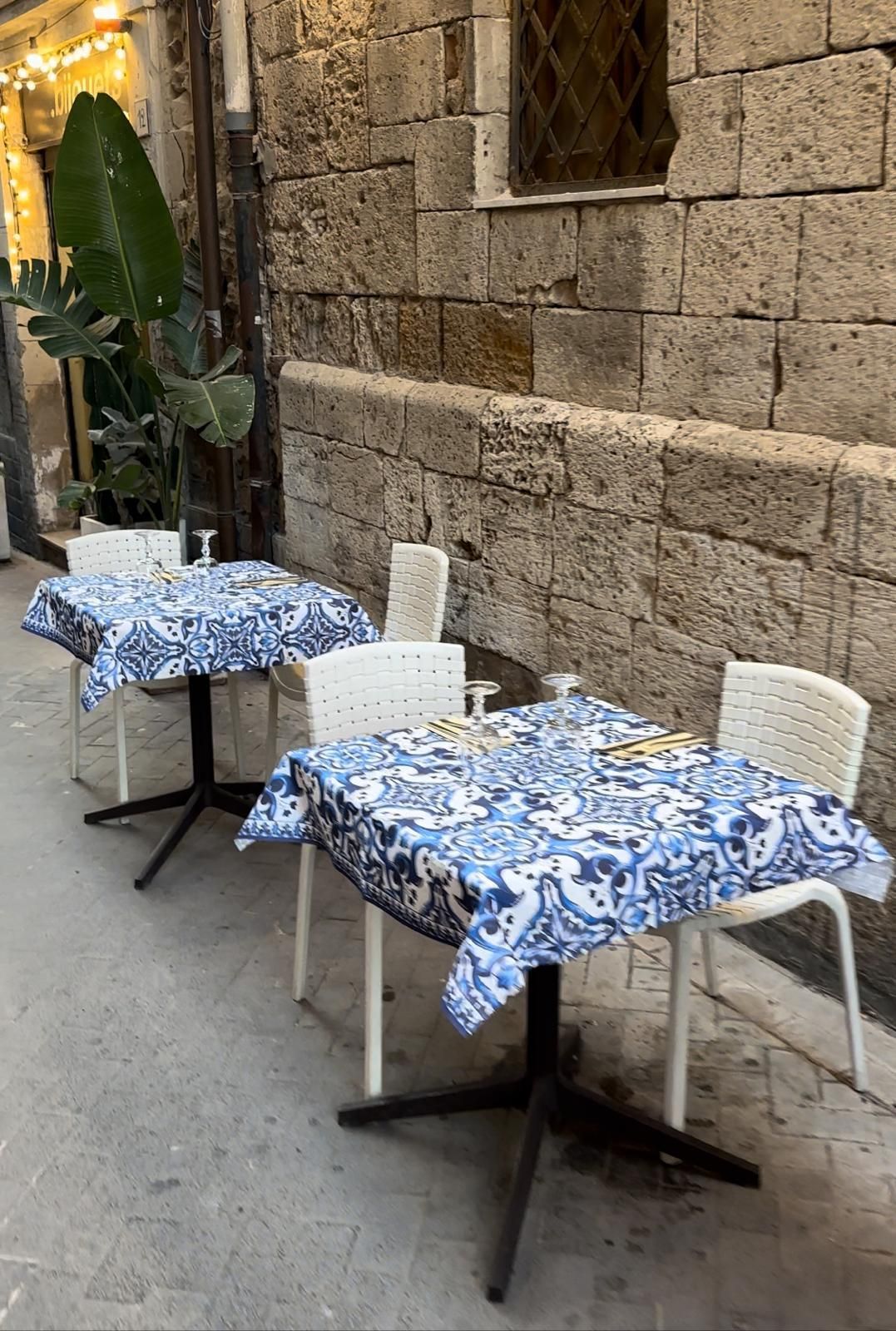 Due tavoli con tovaglie blu e bianche e sedie bianche di fronte a un muro di mattoni ristorante siciliano a Siracusa.