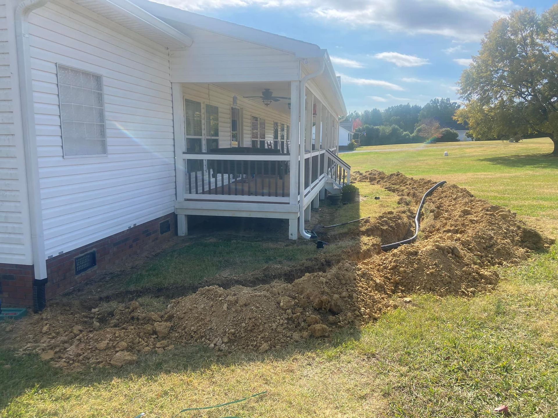 A trench dug along a white house with a porch in a grassy yard, cloudy sky.