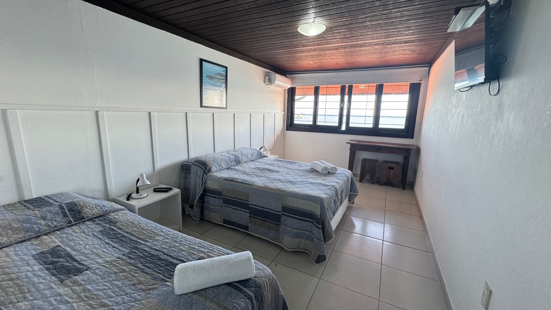 Quarto de hotel com duas camas de solteiro, pequena escrivaninha perto da janela e uma TV na parede.