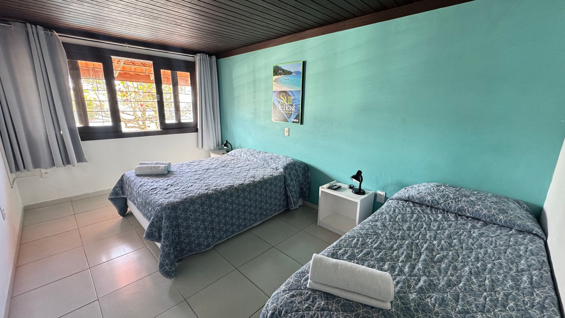 Quarto com duas camas de solteiro, parede turquesa e janela com cortinas.