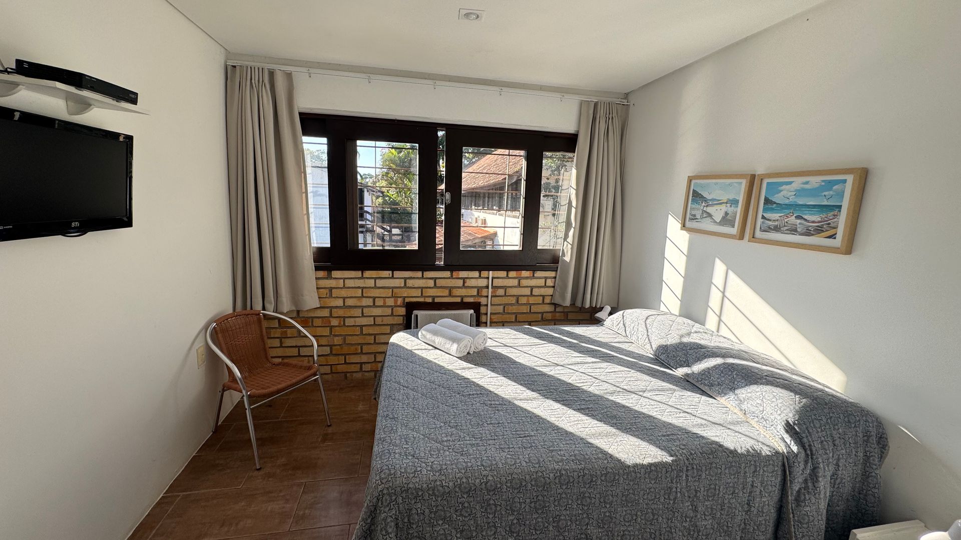 Um pequeno quarto com uma cama, janela e TV. A luz do sol entra.