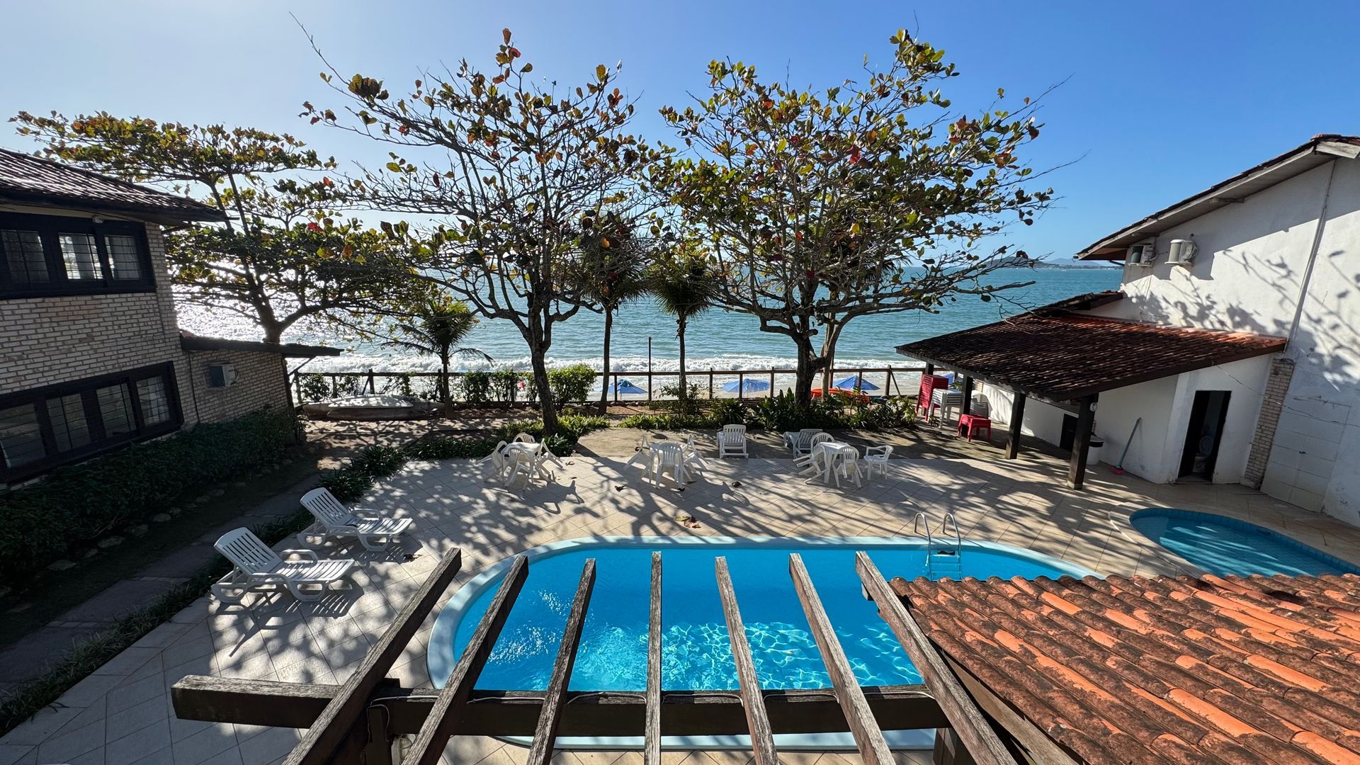 Vista da piscina e da praia de um terraço. Árvores e cadeiras cercam a piscina; o oceano ao fundo.