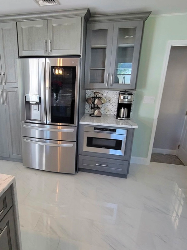 Stainless steel refrigerator and built-in microwave flanked by grey cabinets. A countertop with coffee maker and accessories is on the right.