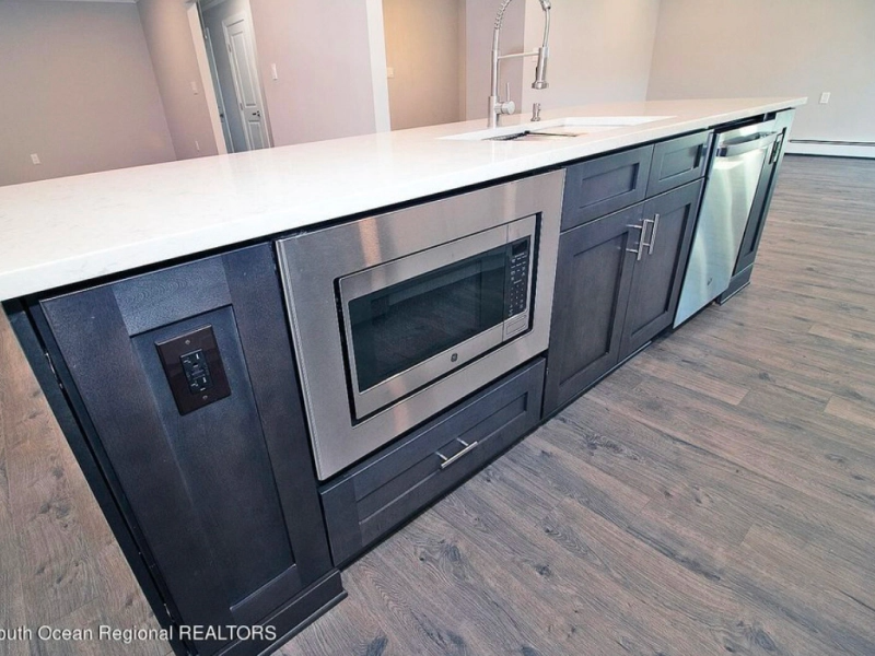 Kitchen island with dark gray cabinets, stainless steel microwave and dishwasher, white countertop, and gray wood flooring.
