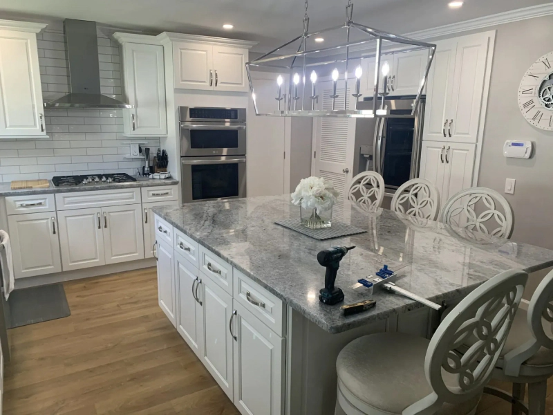 White kitchen with large island, cabinets, and appliances. Stainless steel oven, range hood, and chandelier. Bar stools.