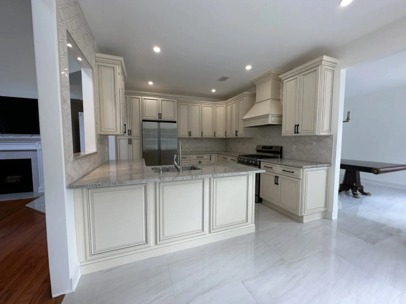 Elegant, cream-colored kitchen with a stainless steel refrigerator, island, and tiled backsplash.  A dining room with a wooden table is visible in the background.