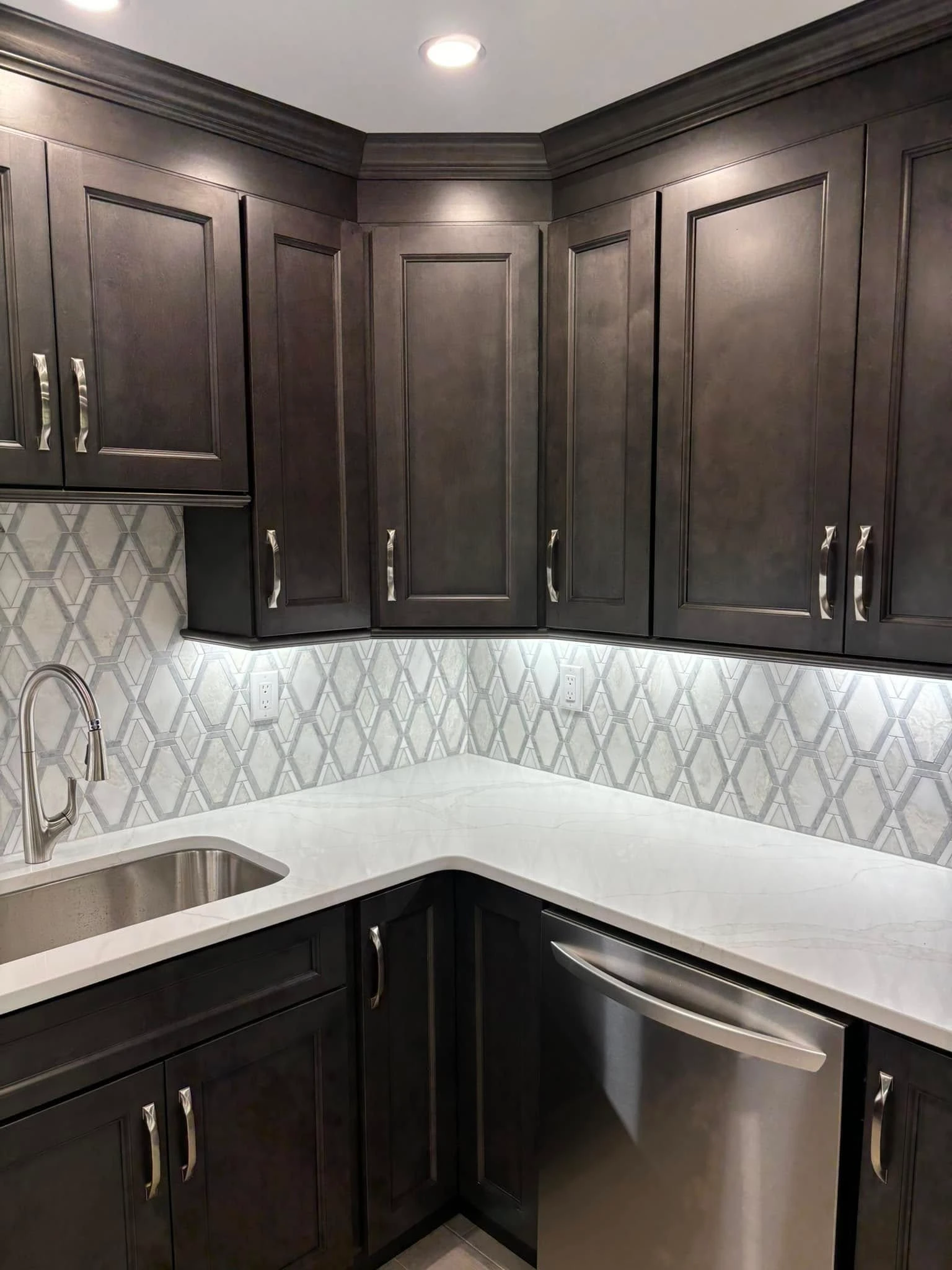Dark gray kitchen cabinets with a stainless steel sink and dishwasher, white countertops, and patterned backsplash tile.