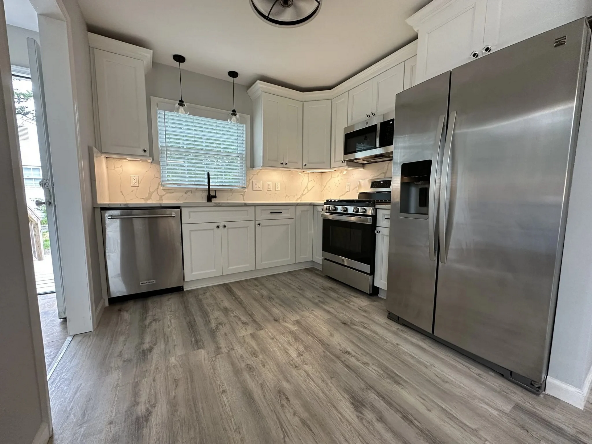 A modern kitchen with white cabinets, stainless steel appliances, and gray flooring. A window and doorway provide natural light.