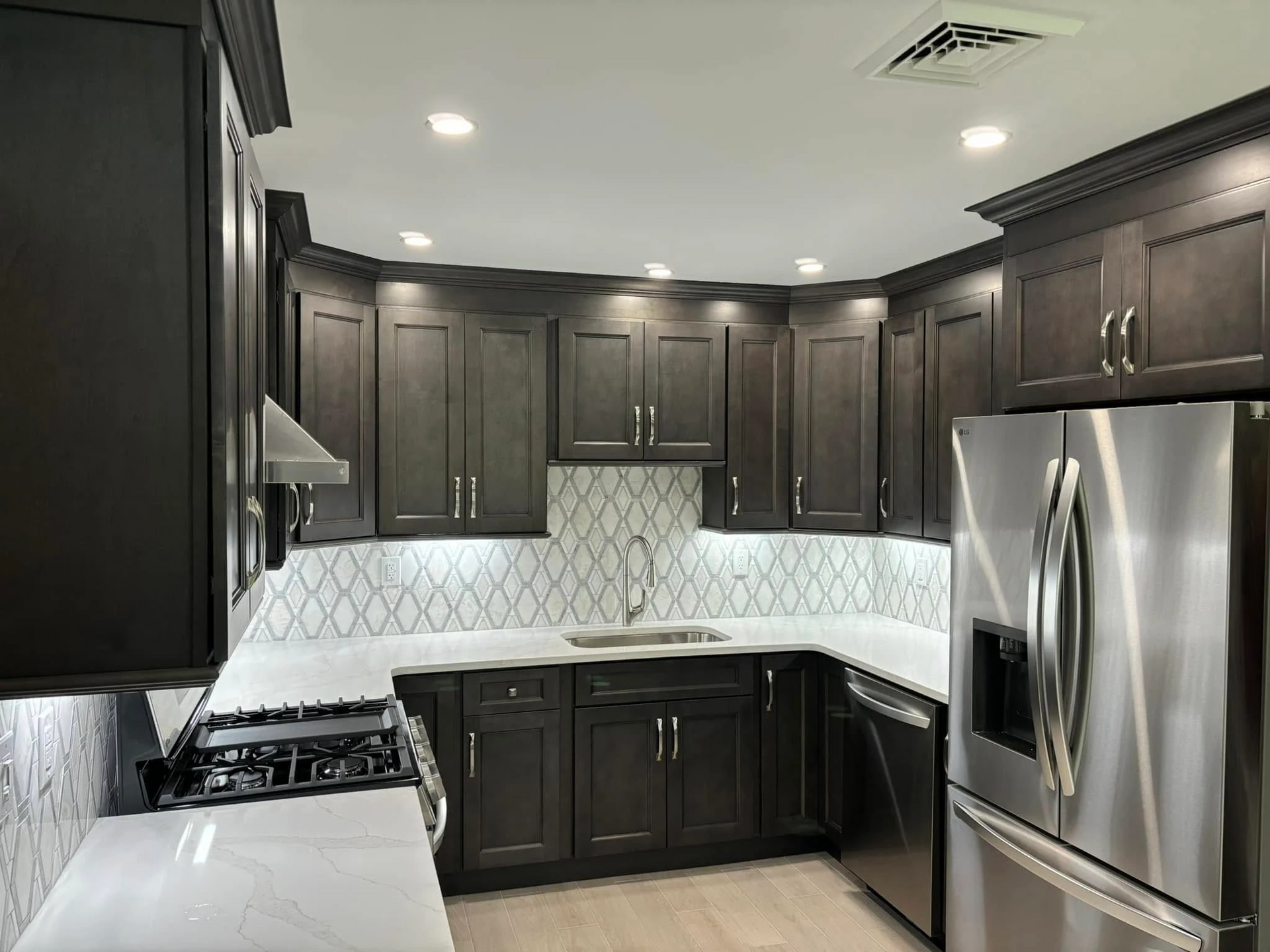 Dark gray kitchen with white countertops, geometric backsplash, and stainless steel appliances. Recessed lighting illuminates the cabinets and workspace.