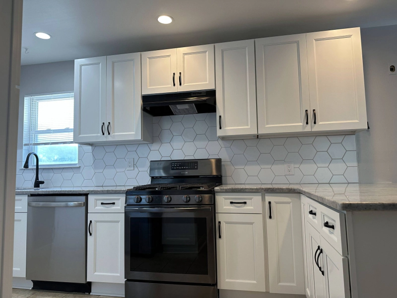 White kitchen with upper and lower cabinets, a stainless steel dishwasher, and a black oven. Hexagonal backsplash and light countertop.