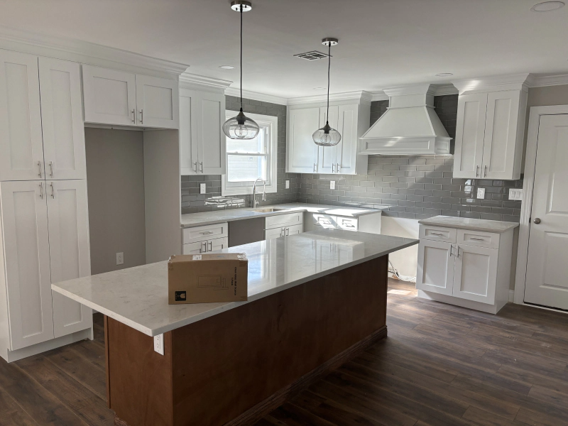 A bright white kitchen with a large island. Features white cabinets, grey tile backsplash, and dark wood island base.