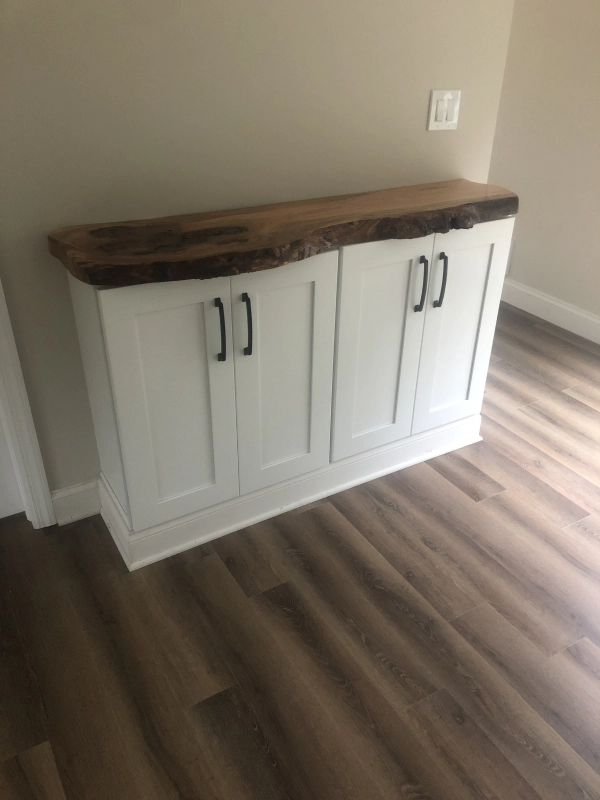 White cabinet with black handles and a live-edge wood top, sitting on a wood floor. A light switch is visible on the wall.