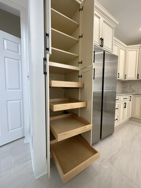 Open pantry cabinet with pull-out drawers and shelves, next to a refrigerator and cream-colored kitchen cabinets.
