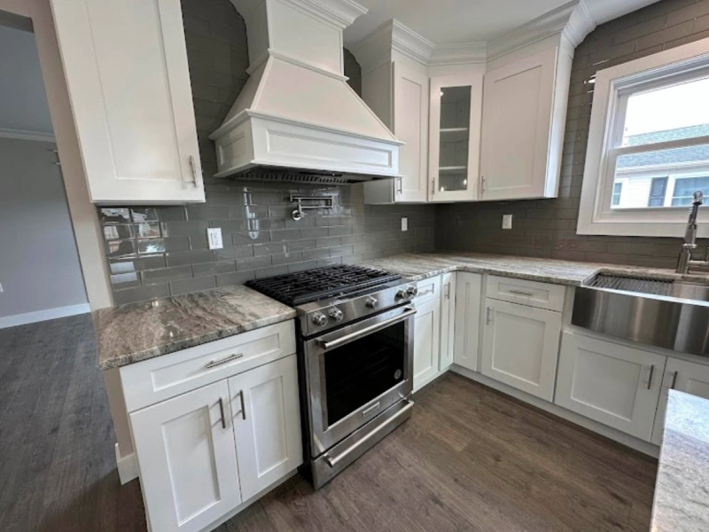A bright, modern kitchen with white cabinets, gray backsplash, and stainless steel appliances. The countertop is light gray.