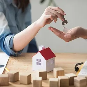 Hands exchanging keys above a small model house with toy blocks on a table