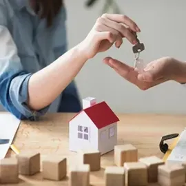 Person placing a house key into an outstretched hand over a model house on a table.