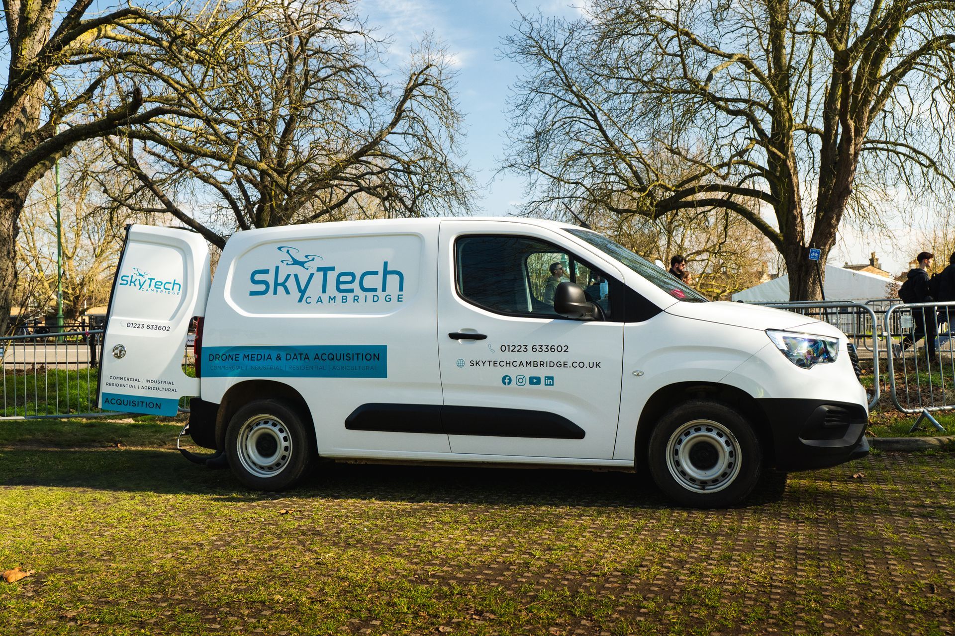 White SkyTech van parked on grass, back door open. Park setting, trees in background.