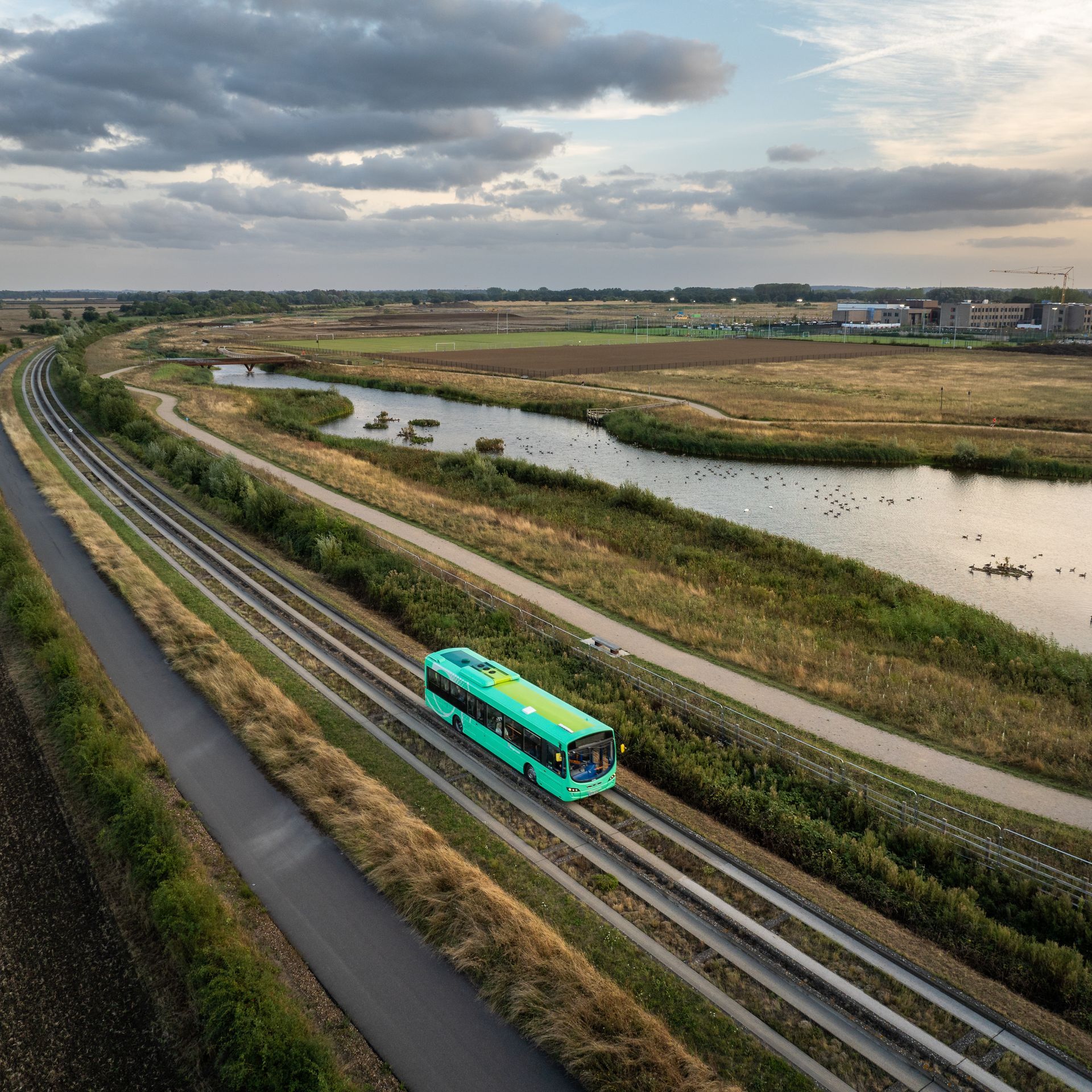 Green bus travels along the Cambridge guided busway tracks through a rural landscape next to Northstowe and its new lakes